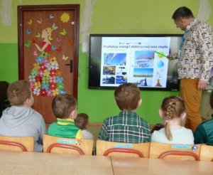 Na zdjęciu Doradca Energetyczny stojący przy wyświetlaczu multimedialnym omawiający sposoby produkcji energii. W tle dzieci w wieku przedszkolnym siedzące i słuchające Doradcę.